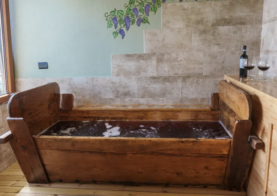 La tinozza del Bagno di Bacco colma di vino rosso, con una bottiglia e due calici sul ripiano accanto